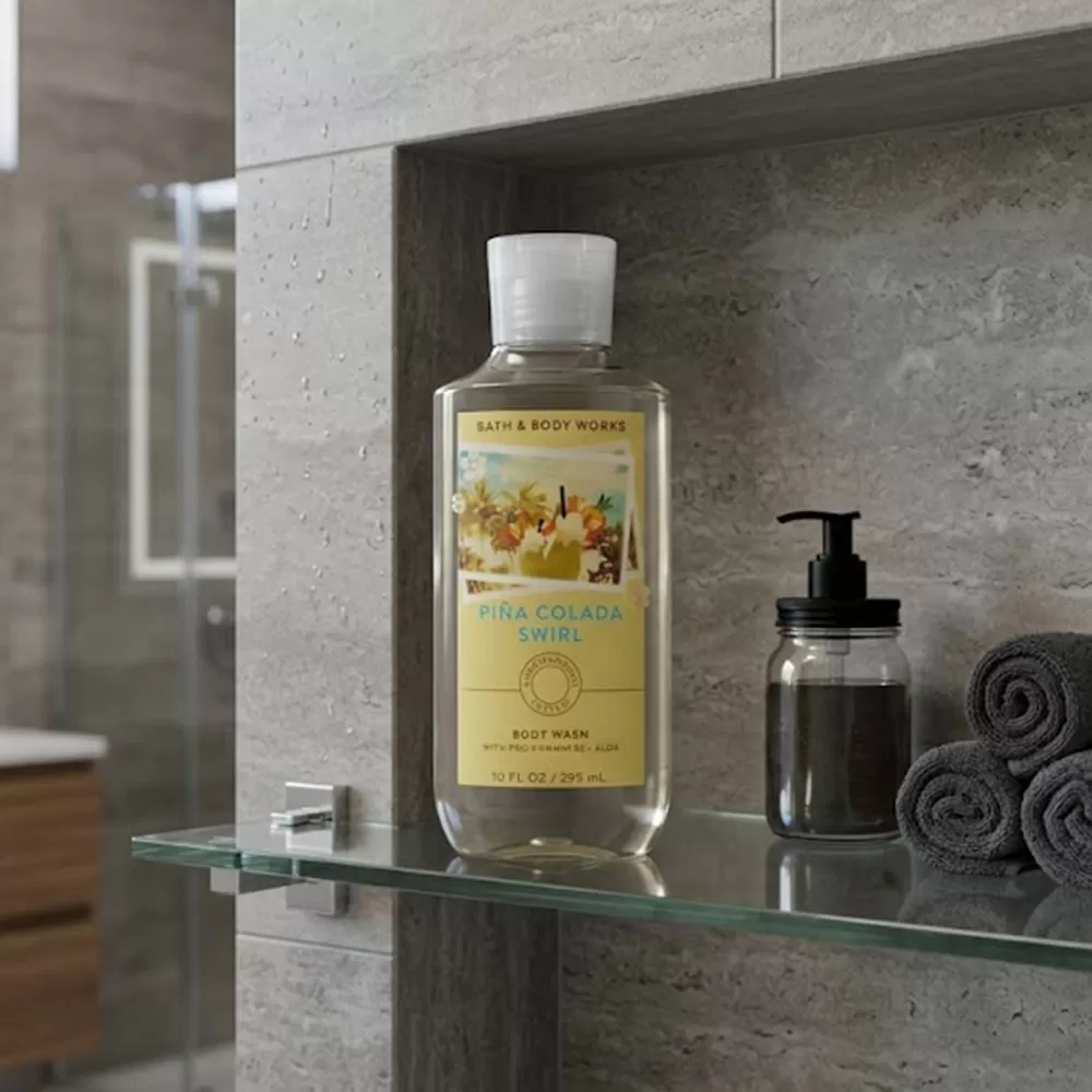 Sữa tắm unisex Body Wash | Bath & Body Works Piña Colada Swirl - chai sữa tắm Bath & Body Works Piña Colada Swirl cạnh ly nước cam và quả cam, trên bàn ăn gỗ.